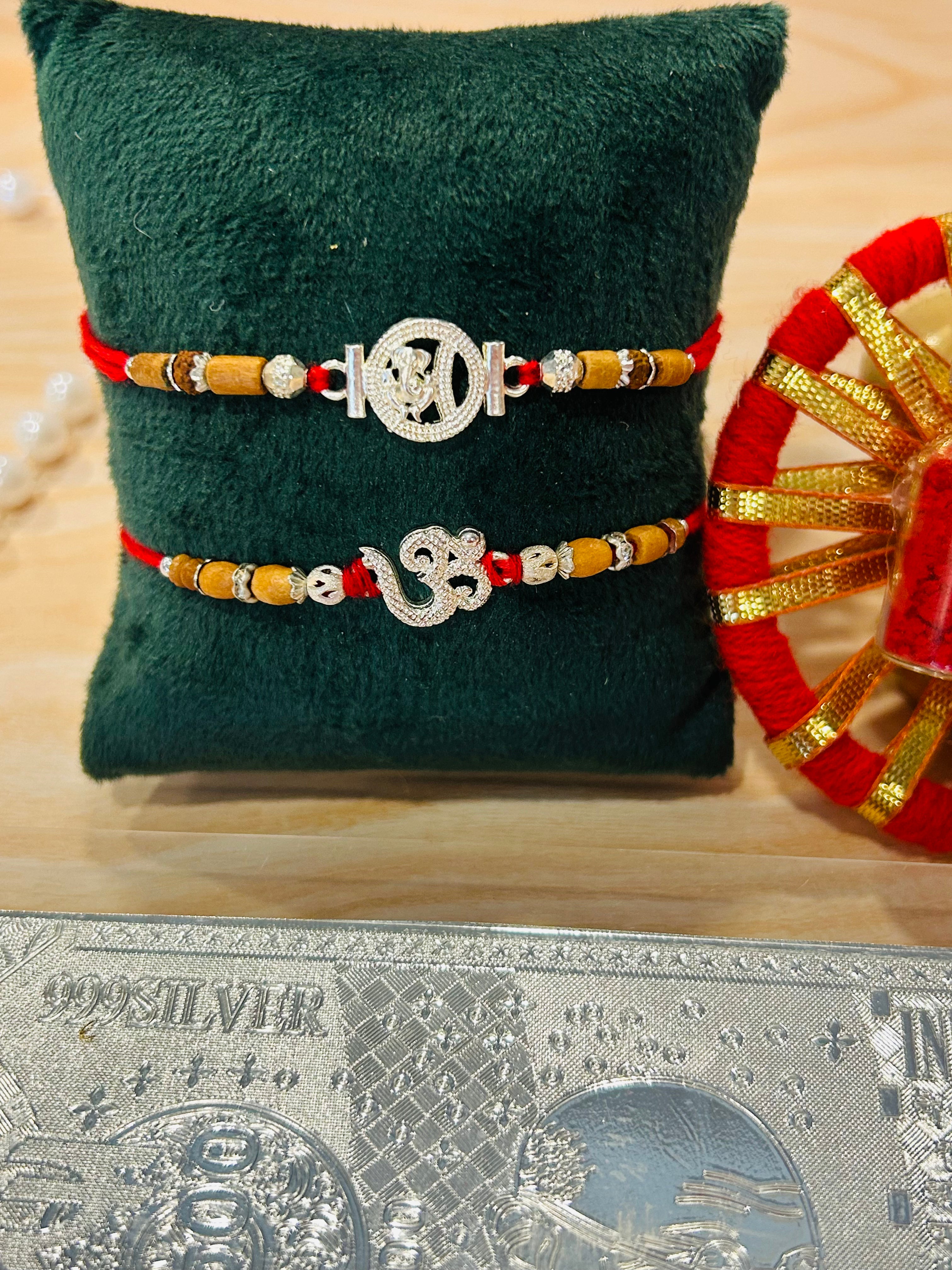Set of 2 silver-finish metal charm rakhis featuring Om and Ganesh motifs with festive threads – by Wrap Shap.