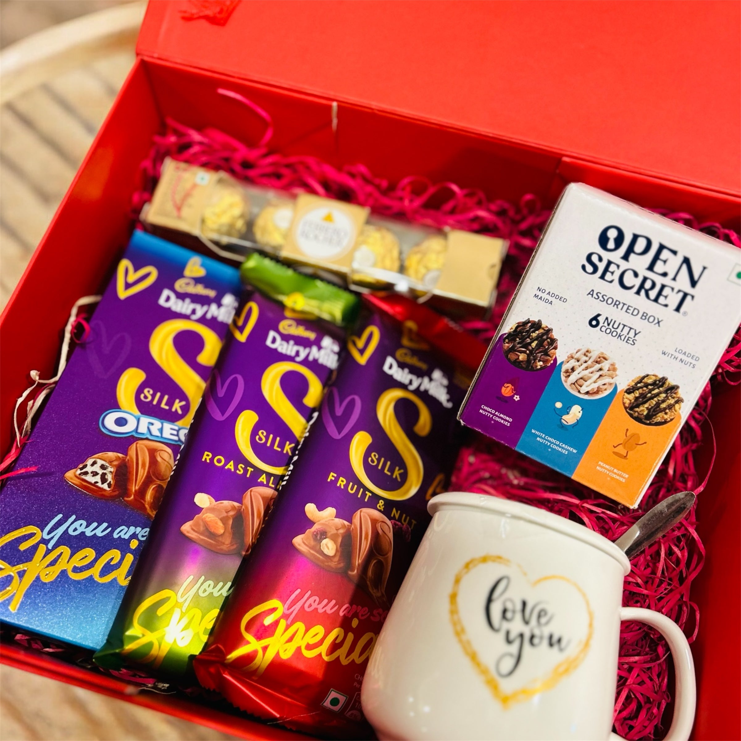 Gift hamper featuring Dairy Milk chocolates, Ferrero Rocher, a 'Love You' coffee mug, and Open Secret assorted cookies in a premium gift box.