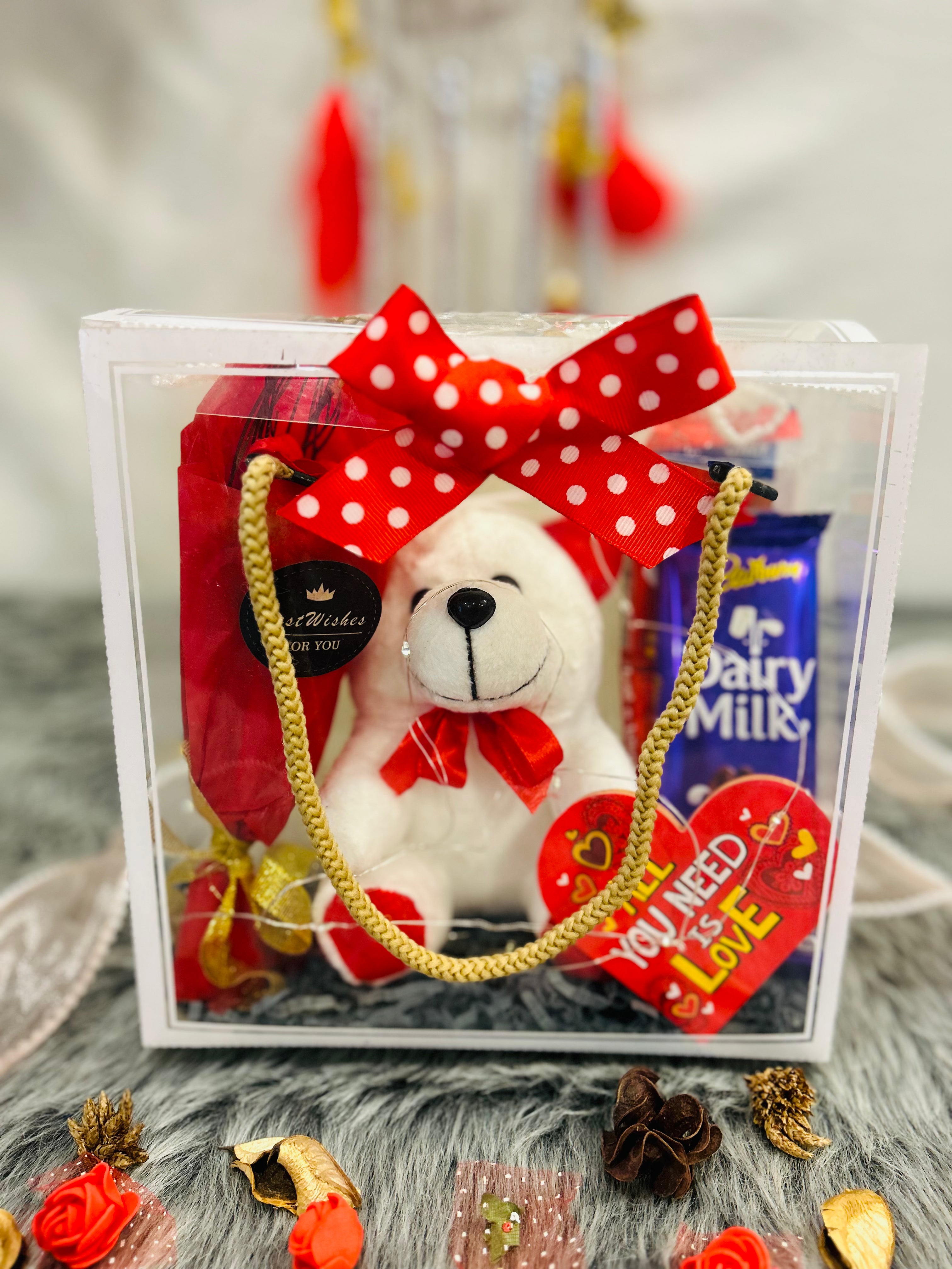PVC transparent hamper featuring a teddy bear, artificial rose bunch, Dairy Milk, Kit Kat chocolates, and a greeting card. Ideal for romantic gifting