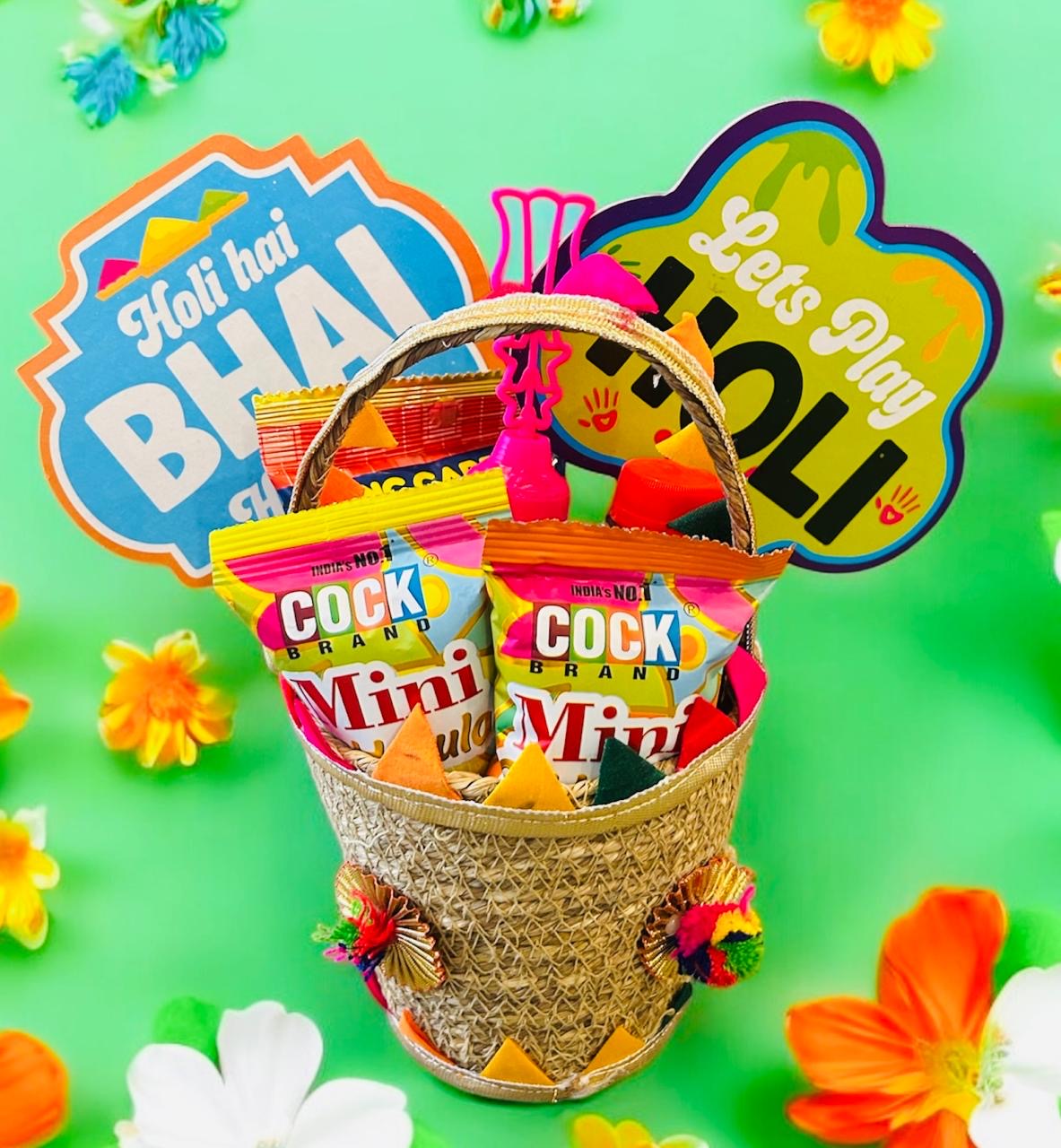 Holi hamper with mini gulal, Tong Garden party snack, Coke, and decorative pichkari in a colorful gift basket.