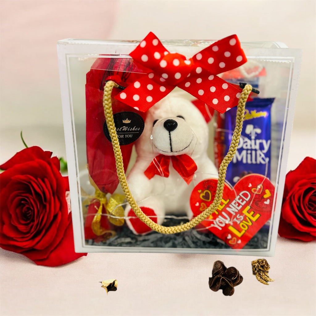 PVC transparent hamper featuring a teddy bear, artificial rose bunch, Dairy Milk, Kit Kat chocolates, and a greeting card. Ideal for romantic gifting.