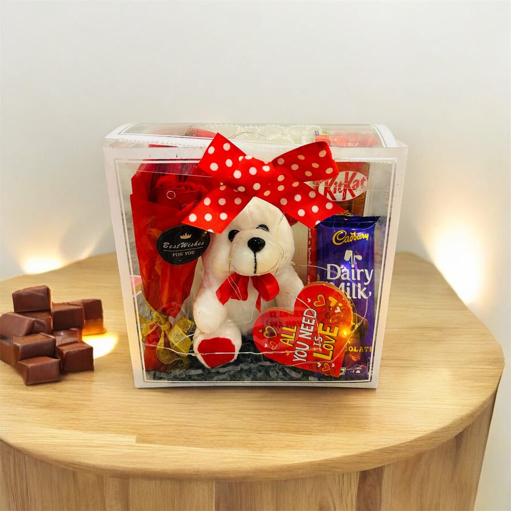 PVC transparent hamper featuring a teddy bear, artificial rose bunch, Dairy Milk, Kit Kat chocolates, and a greeting card. Ideal for romantic gifting.