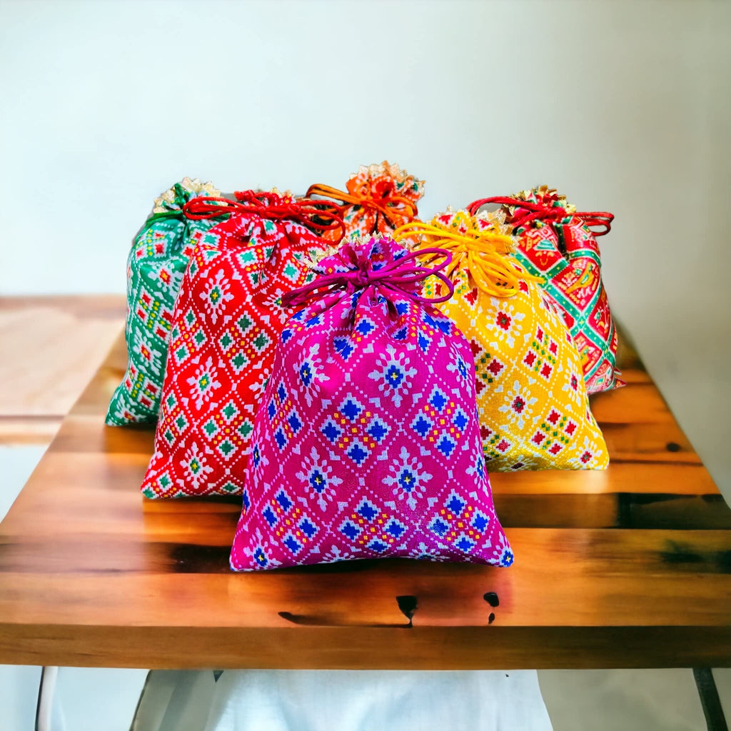 A collection of colorful Patola print potli batwa bags with drawstring closures, displayed on a wooden table.