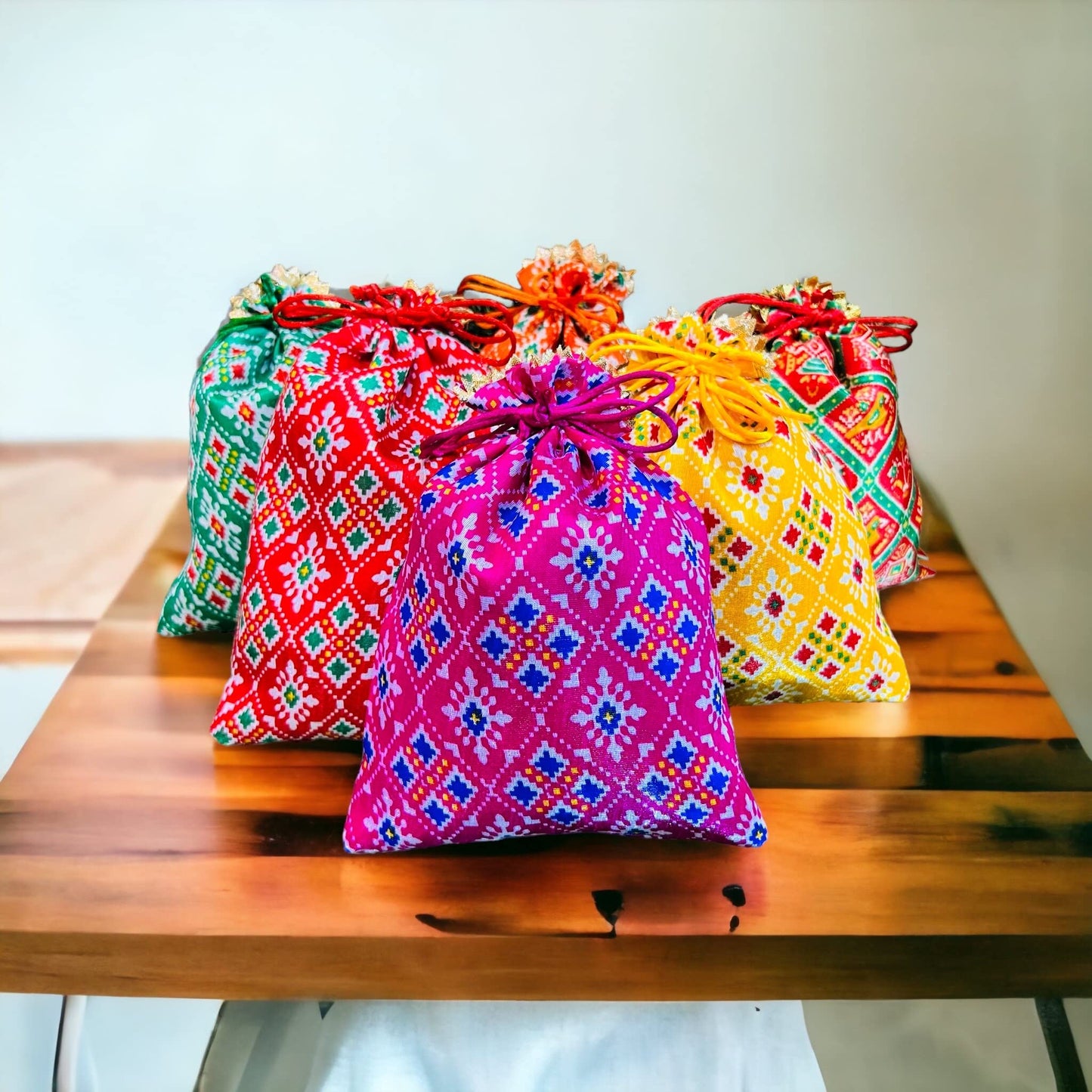 A collection of colorful Patola print potli batwa bags with drawstring closures, displayed on a wooden table.