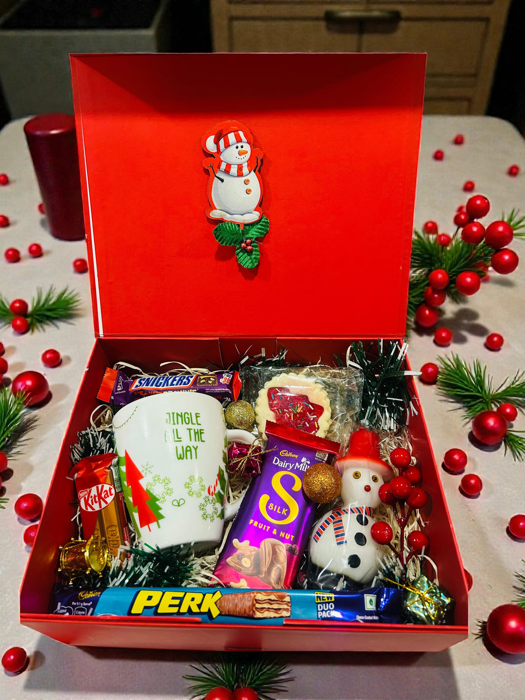 Christmas hamper featuring Christmas-themed mug, candle, cookies and chocolates in red gift box.