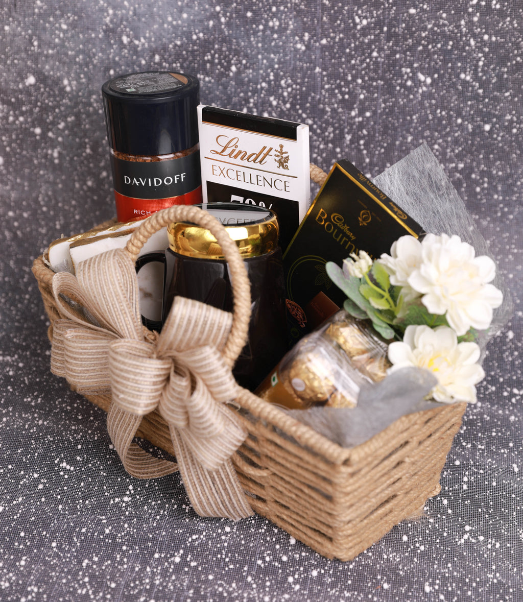 A wicker gift basket filled with assorted treats, including a Davidoff cigarette holder, Lindt Excellence chocolate bar, Raffaello chocolate, and a black mug, decorated with a beige ribbon and white flowers, set against a sparkly gray background.