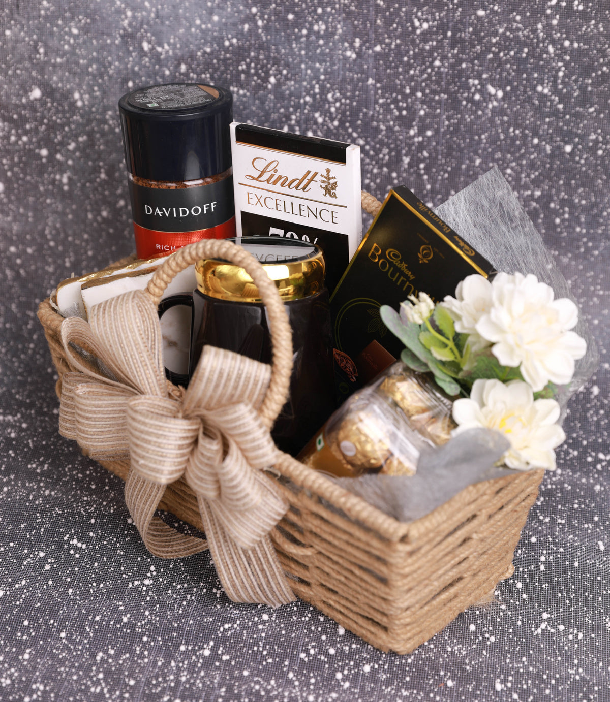 A wicker gift basket filled with assorted treats, including a Davidoff cigarette holder, Lindt Excellence chocolate bar, Raffaello chocolate, and a black mug, decorated with a beige ribbon and white flowers, set against a sparkly gray background.