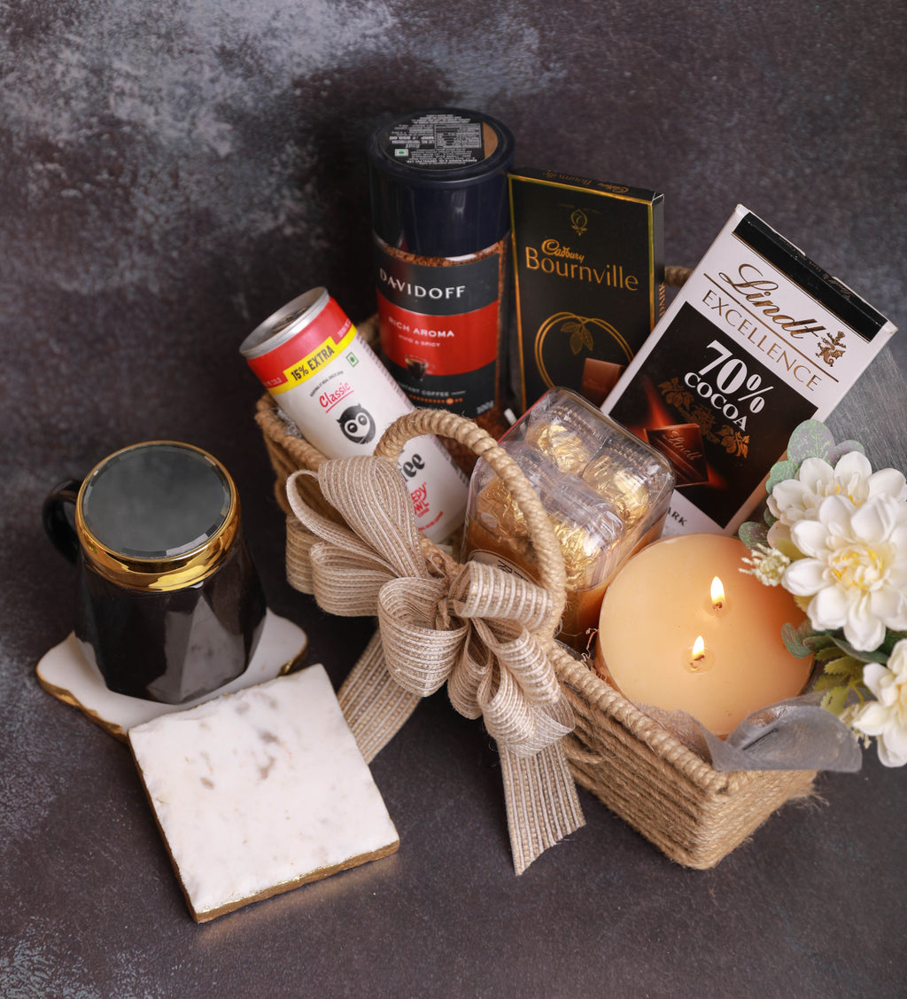 A curated gift basket arrangement on a dark textured surface, featuring a woven basket with a beige ribbon, a black mug with gold trim, a white coaster, a lit orange candle, white flowers, gold leaf in a clear container, a can of coffee, a Cadbury Bournville chocolate bar, and a Lindt 70% cocoa chocolate package.