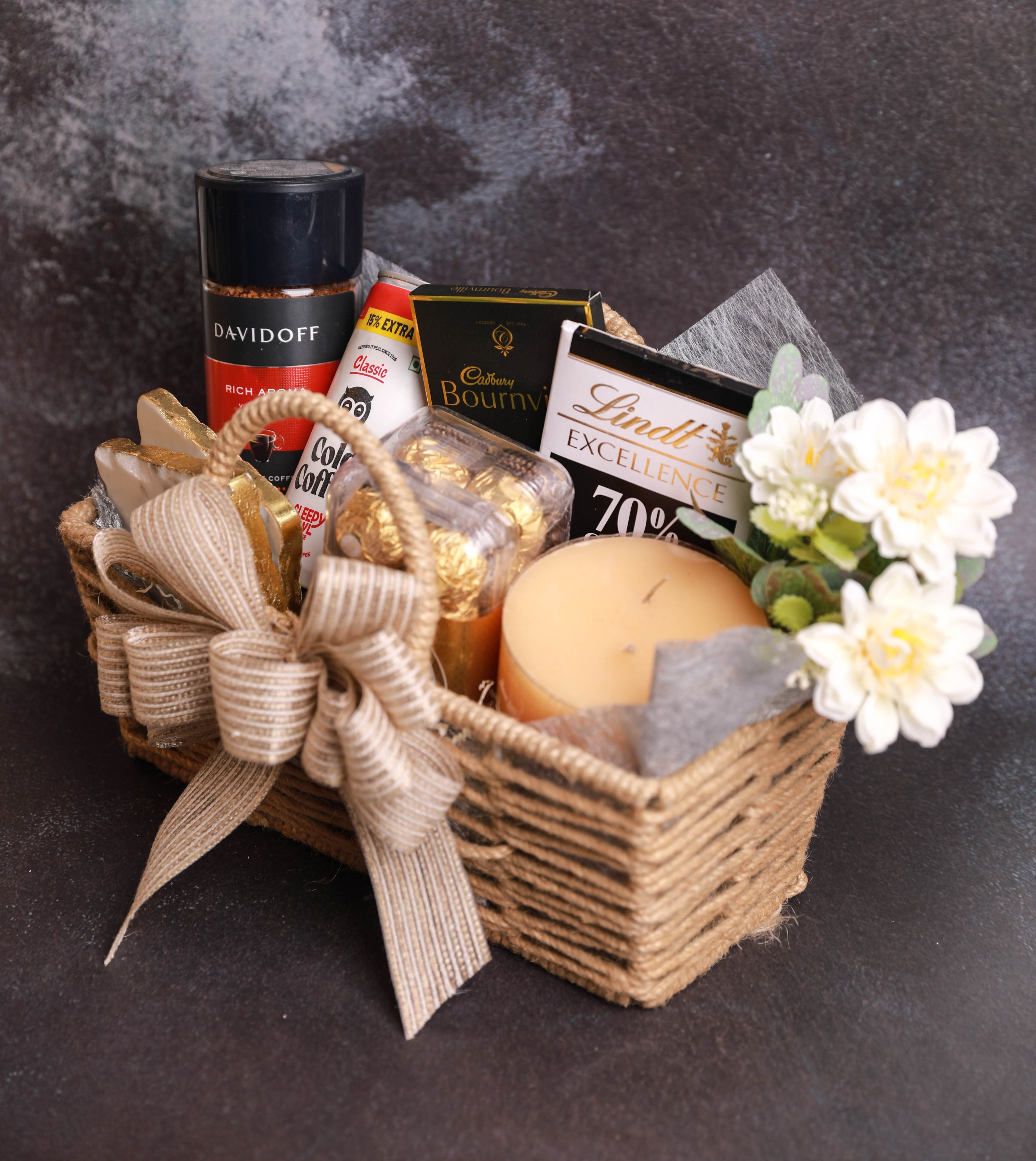A gourmet gift basket filled with assorted treats, including a Davidoff coffee capsule, Lindt Excellence chocolate bar, Ferrero Rocher, and a candle, decorated with a burlap ribbon and white flowers, set against a dark textured background.
