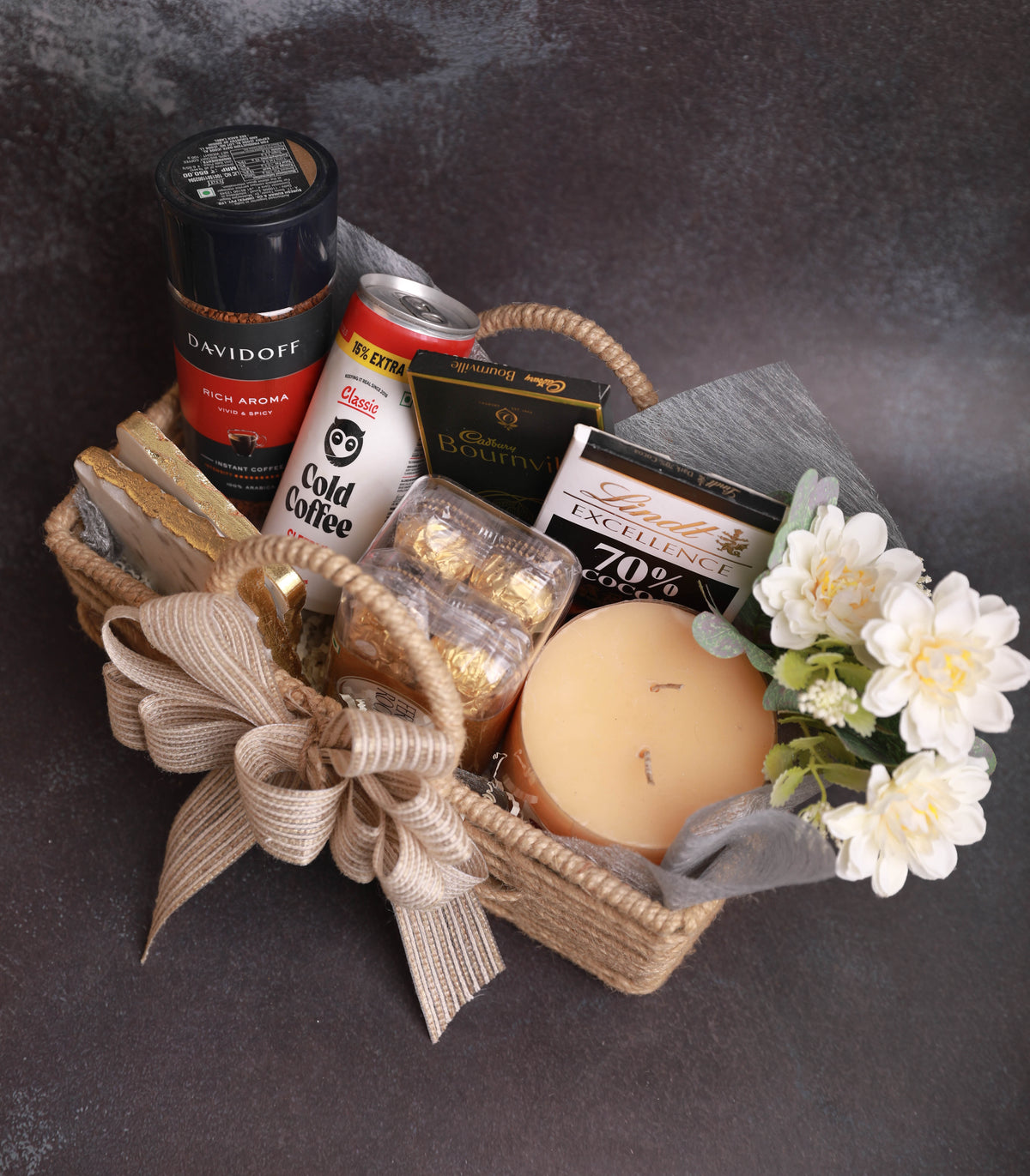 A curated gift basket in a woven natural fiber basket, decorated with a beige ribbon bow. The basket contains a Davidoff Rich Aroma incense stick, a can of Starbucks Cold Coffee, a box of Lindt Excellence chocolate, a candle, gold-leaf wrapped chocolates, and white flowers, all set against a dark textured background.