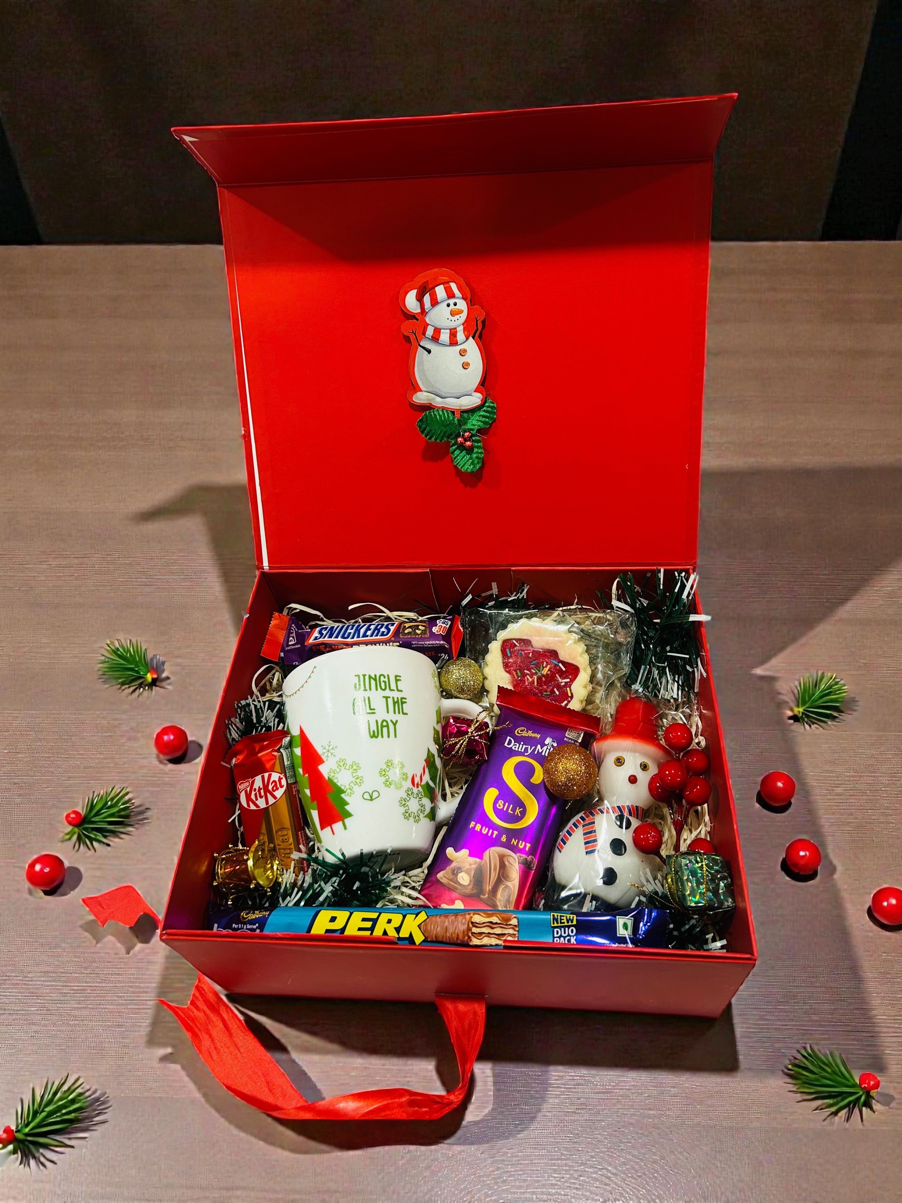 Christmas hamper featuring Christmas-themed mug, candle, cookies and chocolates in red gift box.
