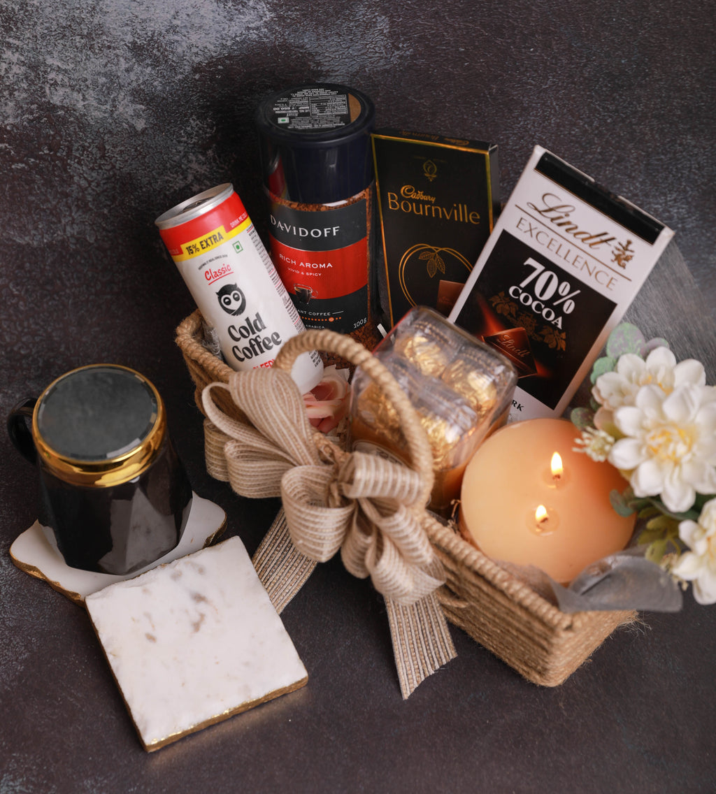 A curated gift basket arrangement on a dark textured surface, featuring a woven basket with a beige ribbon, a lit cream candle, white flowers, a Lindt 70% cocoa box, a Davidoff coffee can, a black coffee mug with gold trim, and a small gold foil packet.
